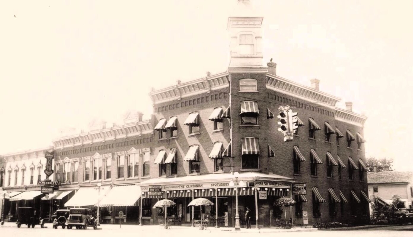 Black-and-white view of the three-story brick Clintonian Hotel in downtown Clinton, Michigan, with striped window awnings, parked automobiles, storefronts and a suspended traffic signal.