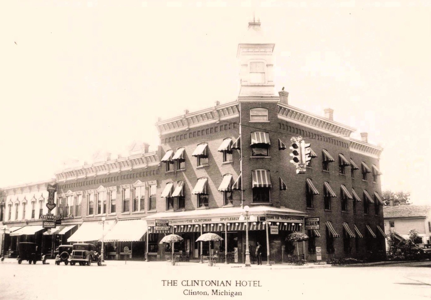 Black-and-white view of the three-story brick Clintonian Hotel in downtown Clinton, Michigan, with striped window awnings, parked automobiles, storefronts and a suspended traffic signal.