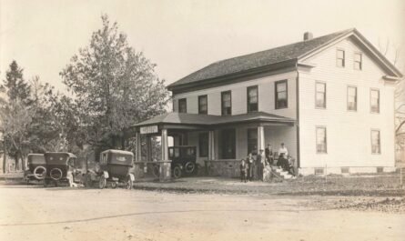 Several people stand outside the two-story Somerset Tavern while three early automobiles are parked beside the porch along an unpaved road.