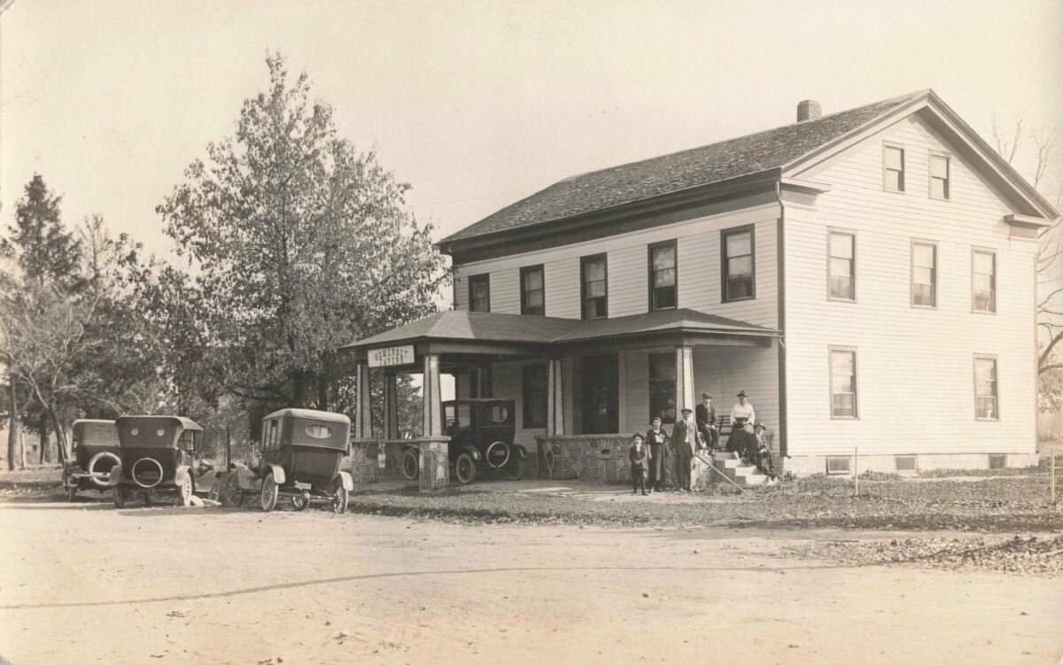 Several people stand outside the two-story Somerset Tavern while three early automobiles are parked beside the porch along an unpaved road.