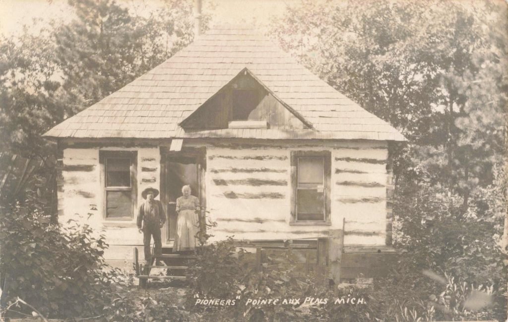 Historic postcard labeled Pioneers showing an unidentified man and woman standing outside a log house at Pointe Aux Pins, Michigan.
