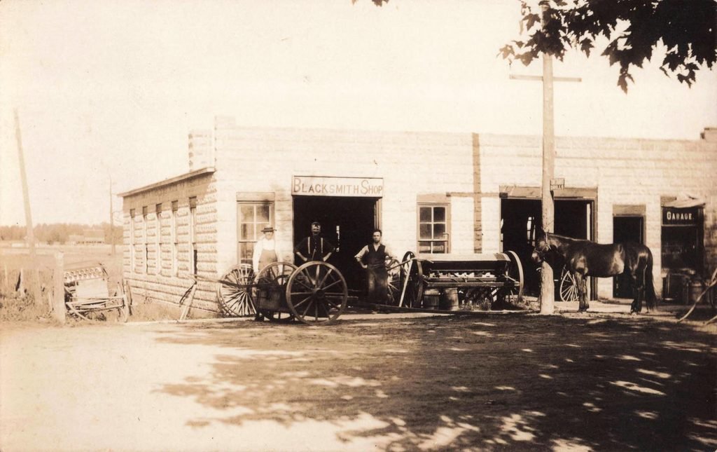 Historic Clifford Michigan blacksmith shop with workers, wagon wheels, farm machinery and horses outside the building.