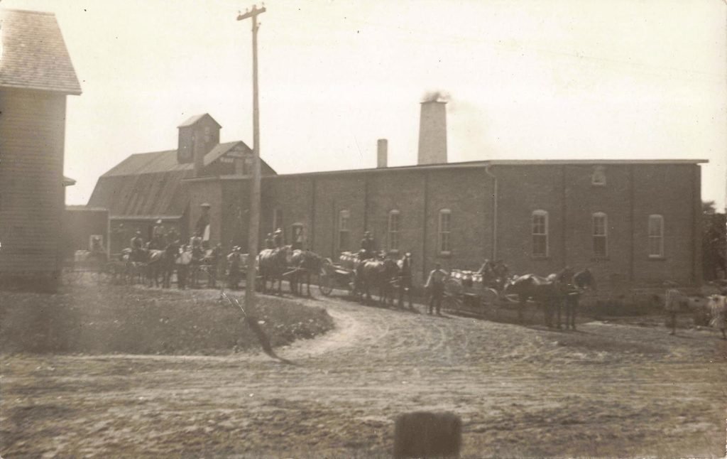 Historic Clifford Michigan creamery with farmers, horse-drawn wagons and a large brick dairy processing building.