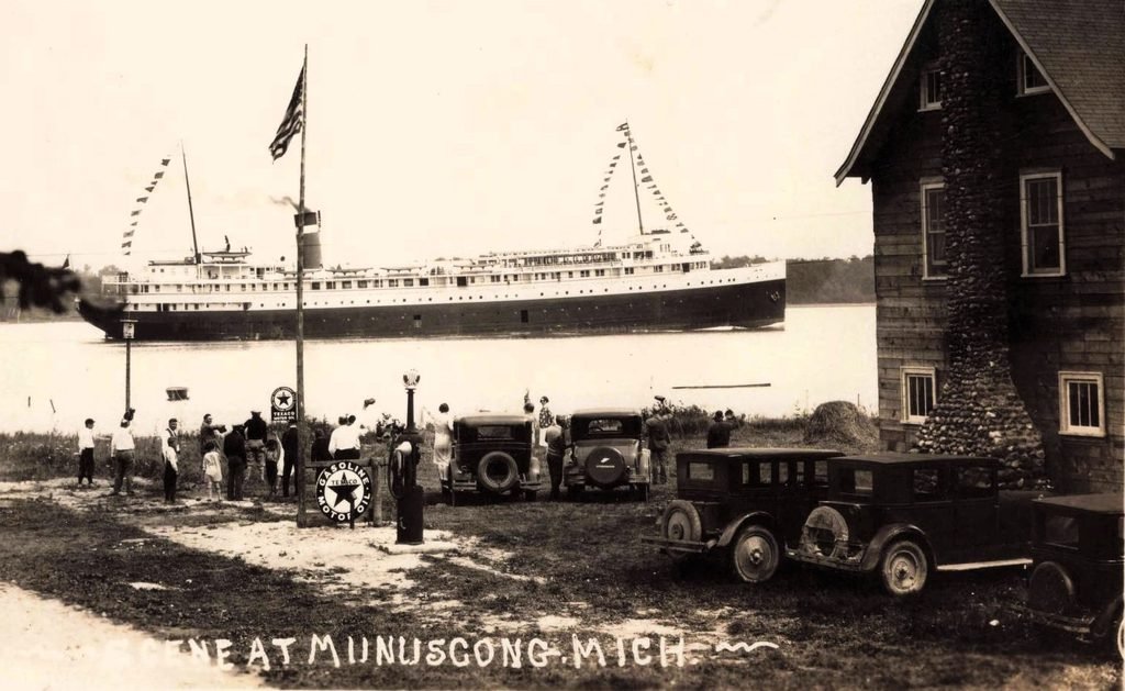 A large passenger steamer passes the inn as people on the bank raise their arms. Its name is uncertain. The scene also shows cars and gasoline pumps beside the lodge. Source: University of Michigan, item 11605.