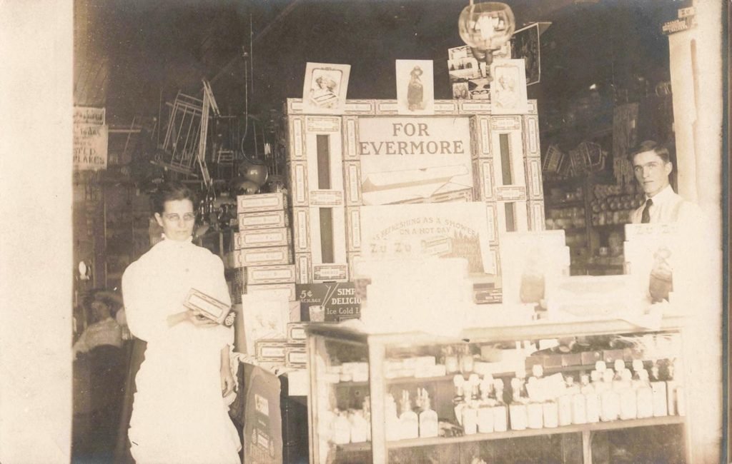 Early Clifford Michigan history -a store with two clerks, a glass display case, bottles, boxes and advertising signs.