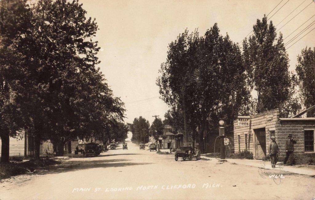 Historic Main Street in Clifford Michigan with early automobiles, a gas pump, Kelly Tires sign and mature street trees.