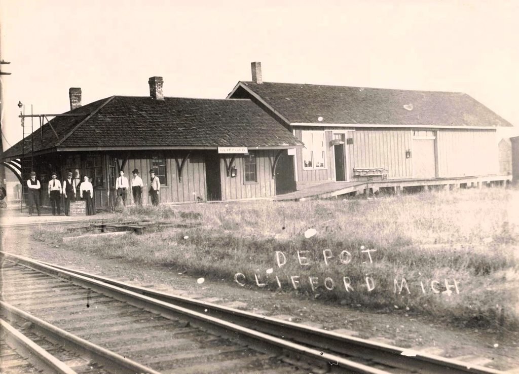 Historic railroad depot in Clifford, Michigan, beside intersecting rail lines with men standing near the station.