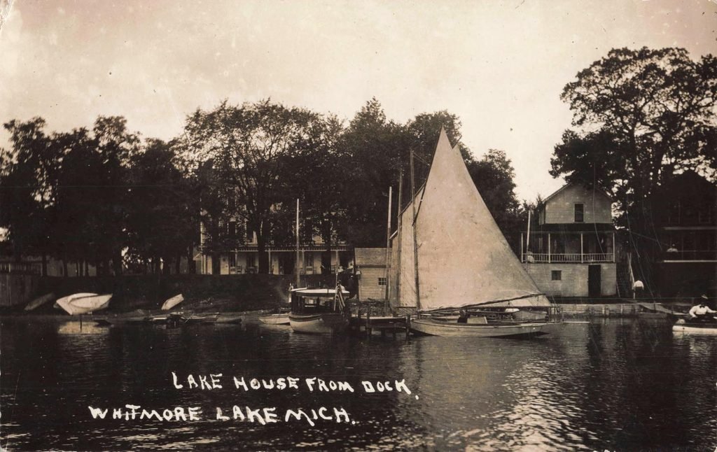 Sailboat on Whitmore Lake, with waterfront resort buildings and smaller boats beyond