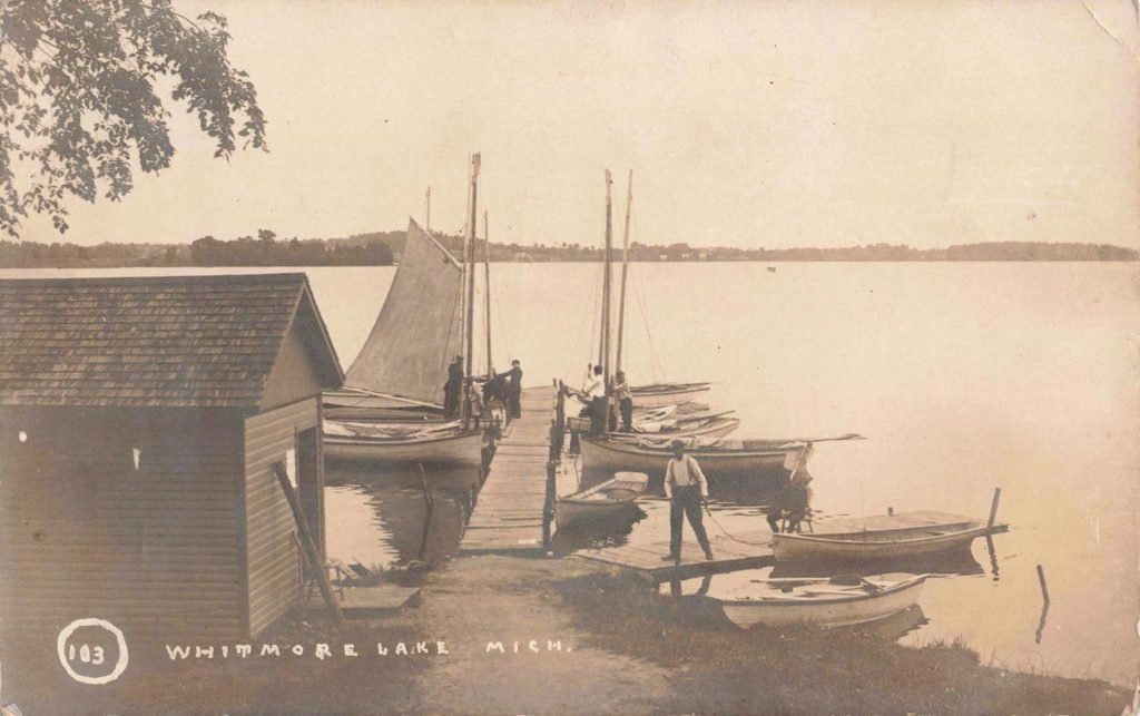 Sailboat on Whitmore Lake, with waterfront resort buildings and smaller boats beyond