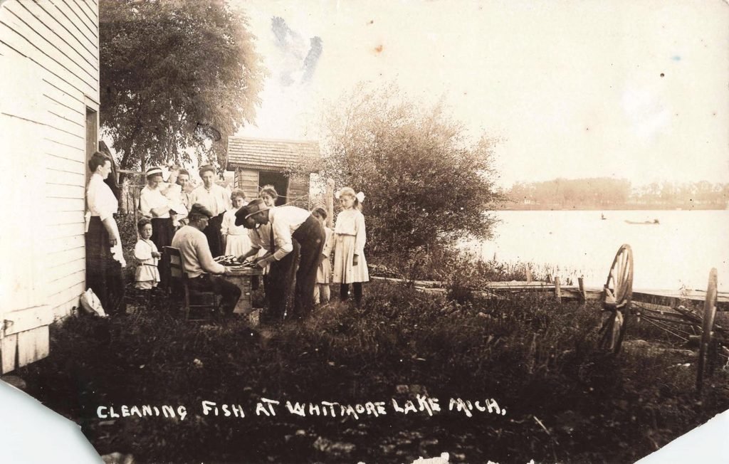 People standing and sitting near the side of a wooden building while cleaning fish beside the lake